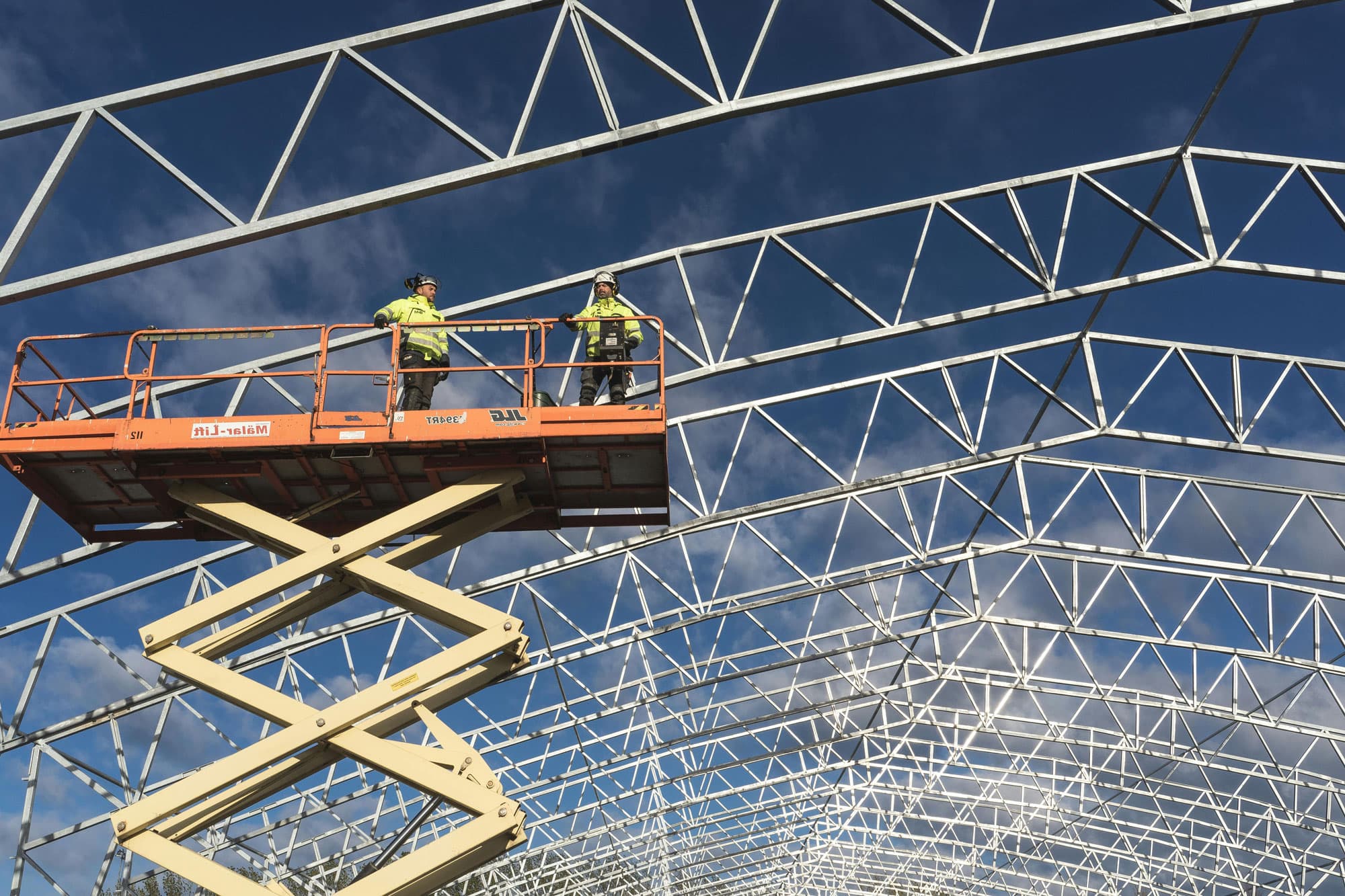 Zwei Bauarbeiter in einer Hebebühne bei Arbeiten an einem Stahlrahmen unter blauem Himmel.