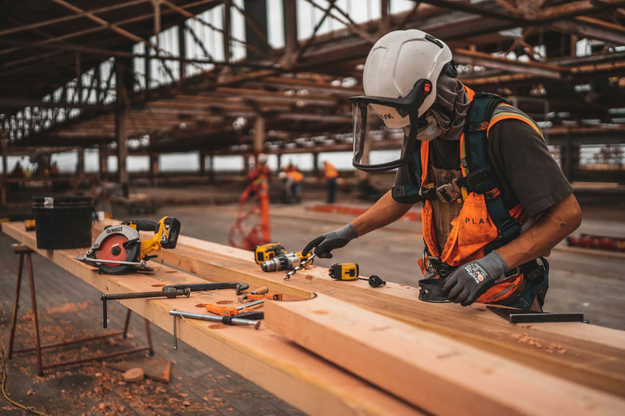 Ein Bauarbeiter mit Schutzhelm und Schutzbrille arbeitet auf einer Baustelle. Er benutzt verschiedene Werkzeuge auf einem Arbeitstisch, um Holz zu bearbeiten.