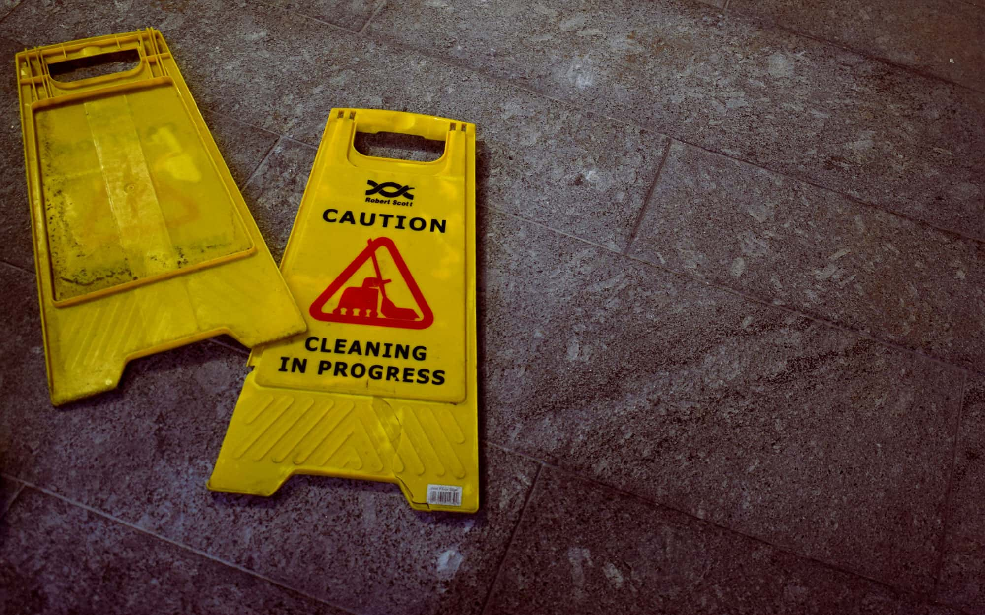 Ein gelbes Warnschild mit der Aufschrift 'Caution: Cleaning in Progress' liegt auf einem gefliesten Boden und warnt vor Reinigungsarbeiten.