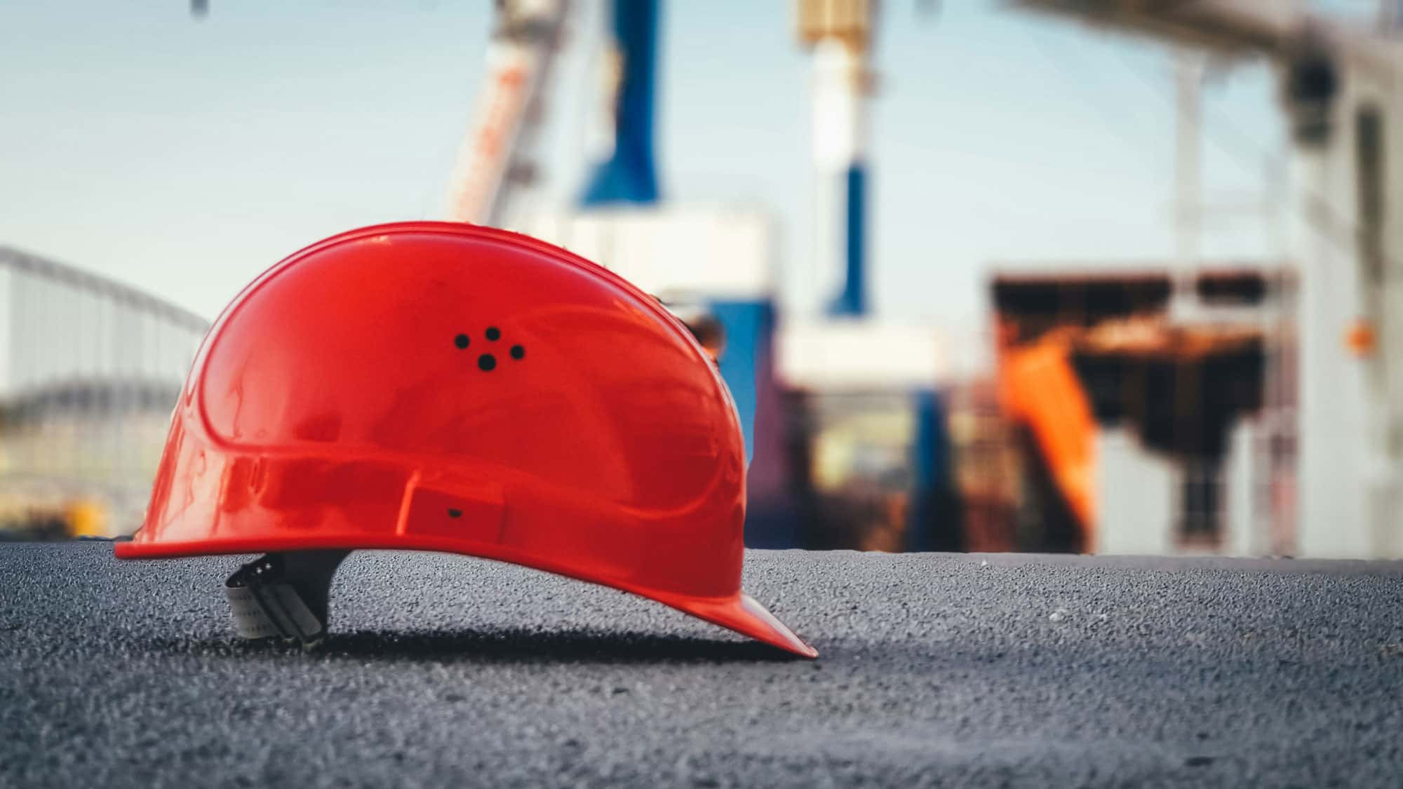 Ein roter Schutzhelm liegt auf dem Boden einer Baustelle. Im Hintergrund sind unscharf Baugerüste und Kräne zu sehen, die eine aktive Bauumgebung andeuten.