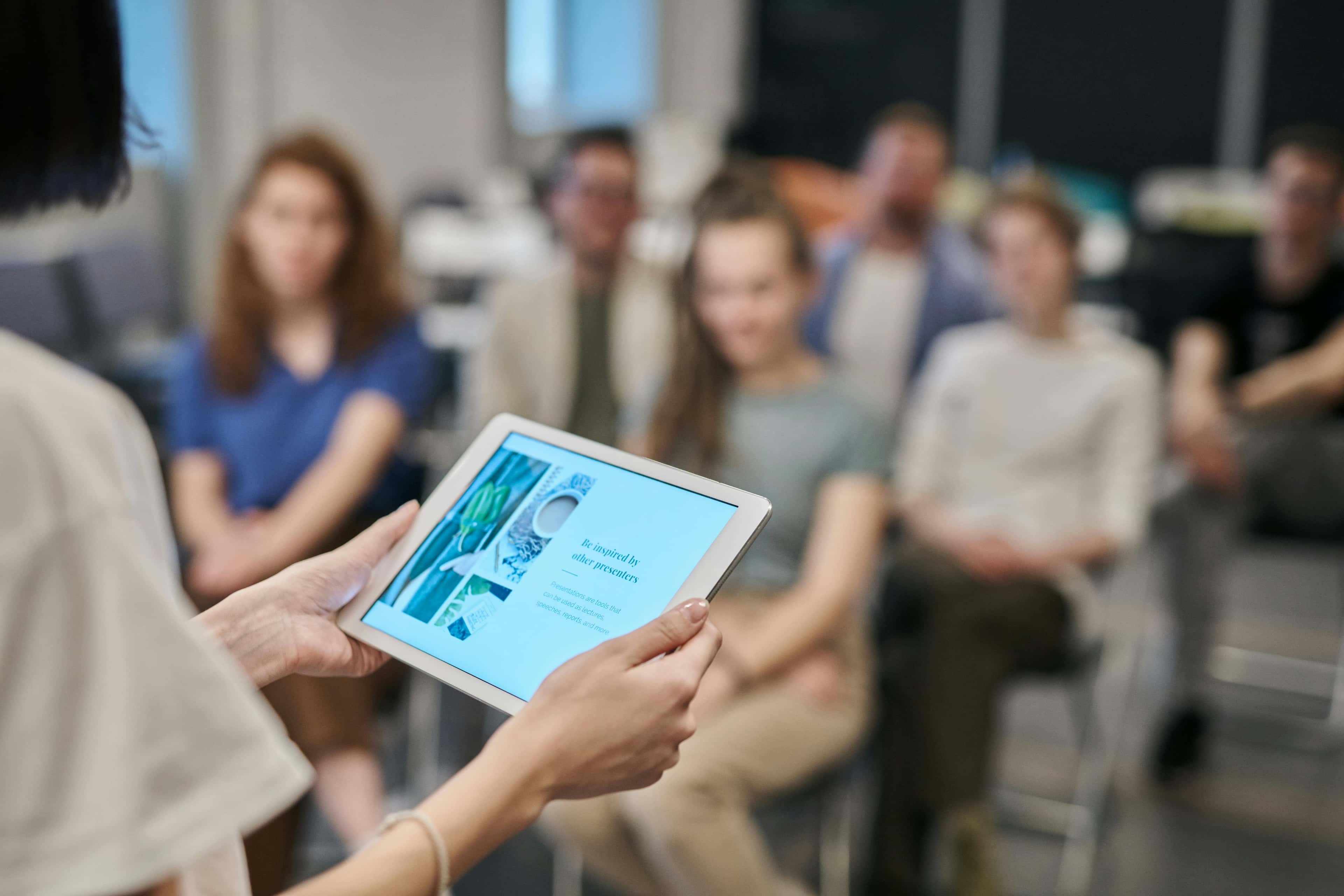 Eine Person hält ein Tablet, das eine Präsentation zeigt, während im Hintergrund eine Gruppe von Menschen unscharf zu sehen ist, die der Präsentation aufmerksam folgt.