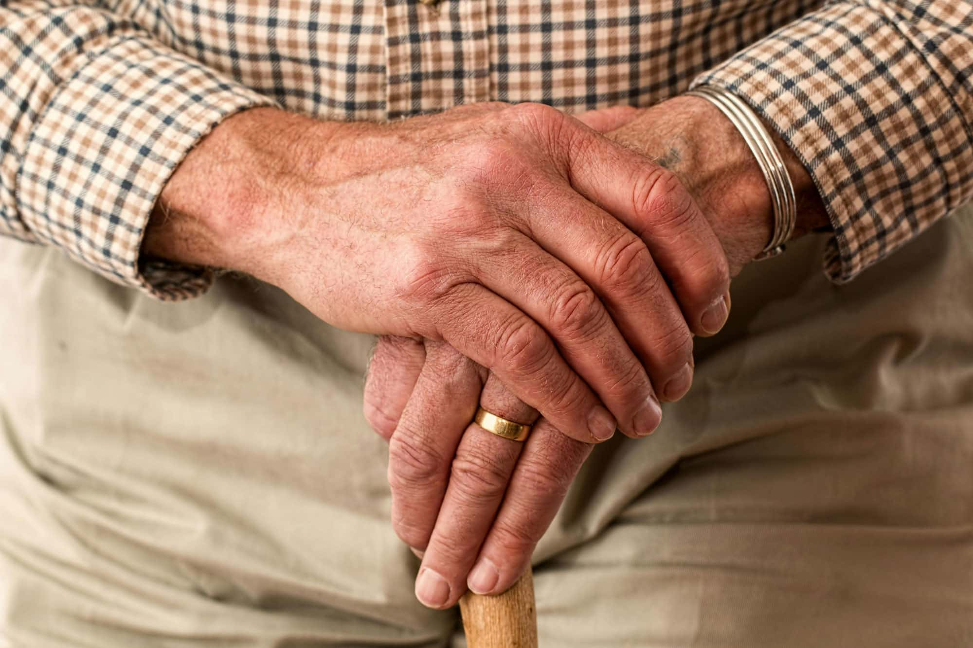 Nahaufnahme der Hände eines älteren Mannes, die auf einem Gehstock ruhen. Der Mann trägt ein kariertes Hemd und einen goldenen Ring am Ringfinger.