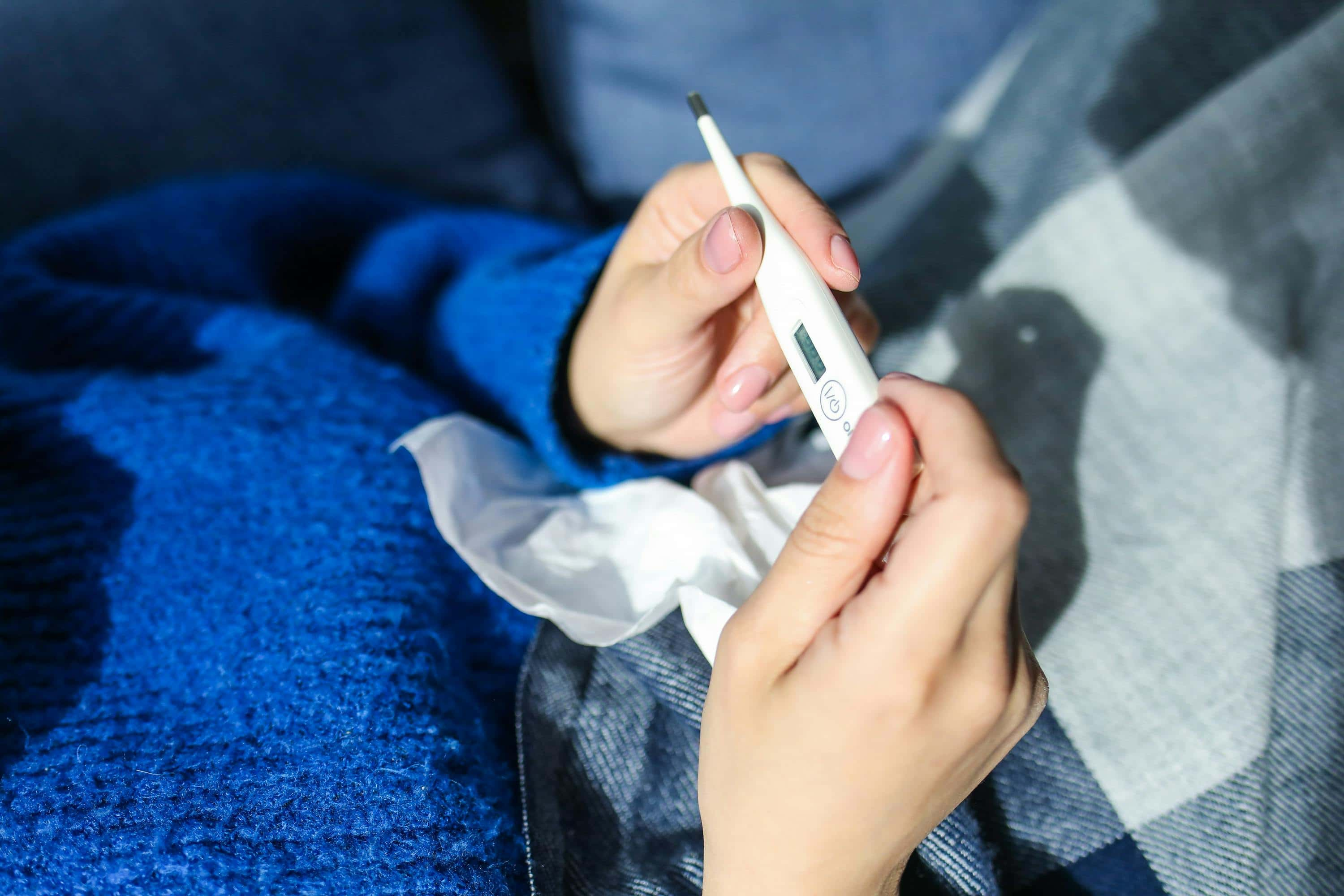 Eine Person in einem blauen Pullover hält ein Thermometer und ein Taschentuch. Die Szene deutet auf eine Gesundheitsüberprüfung hin, möglicherweise wegen Krankheit.