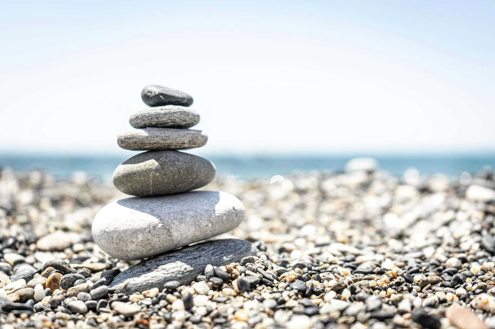 Ein kleiner Stapel von Steinen, sorgfältig aufeinander gestapelt, liegt auf einem kieseligen Strand mit verschwommenem Meereshintergrund. Das Bild symbolisiert Balance und Ruhe in einer natürlichen Umgebung.