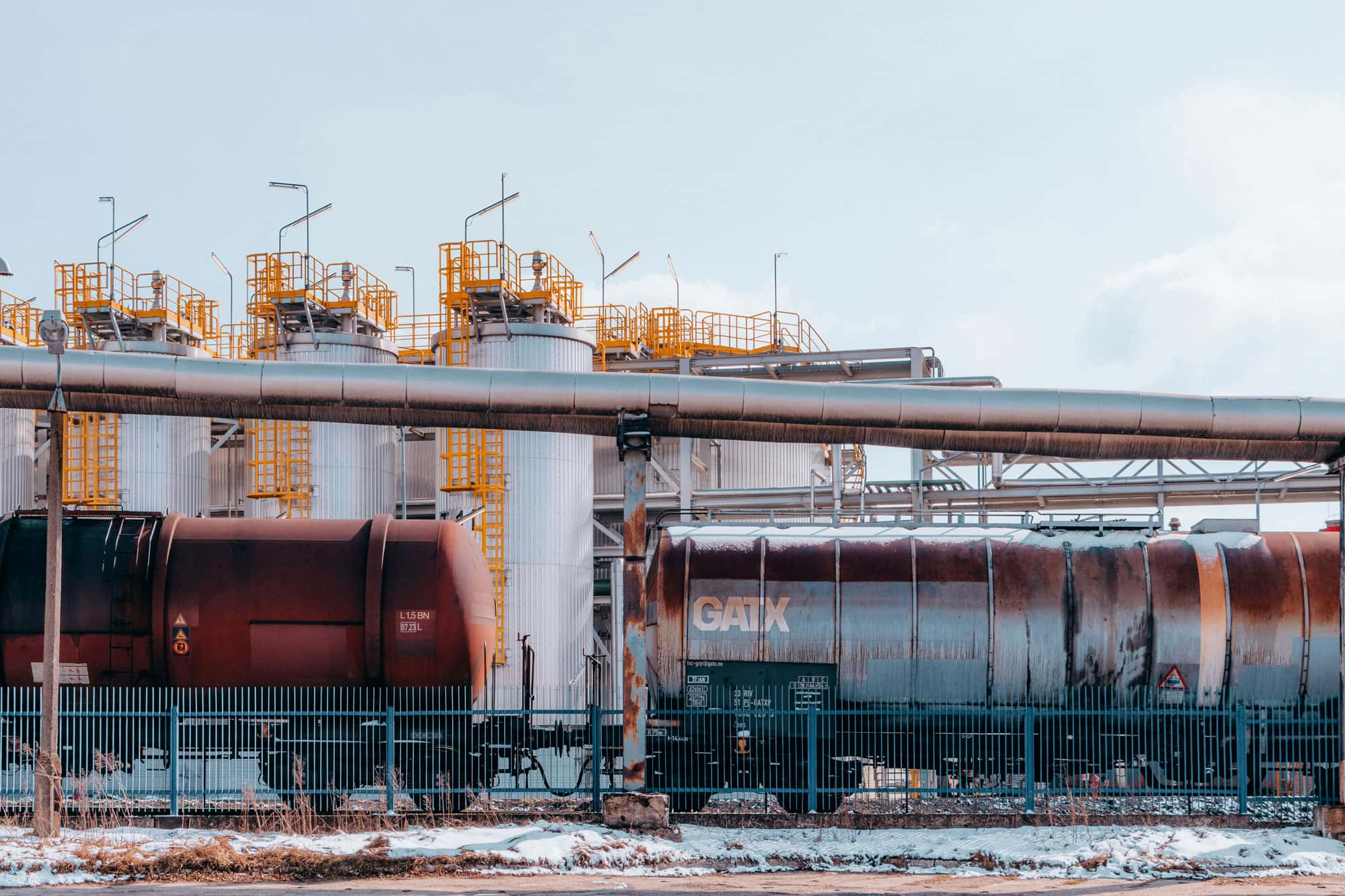 Eine Industrieanlage mit mehreren Lagertanks und Eisenbahnwaggons im Vordergrund. Gelbe Leitern und Plattformen sind an den Tanks angebracht. Ein blauer Zaun verläuft entlang der Anlage.