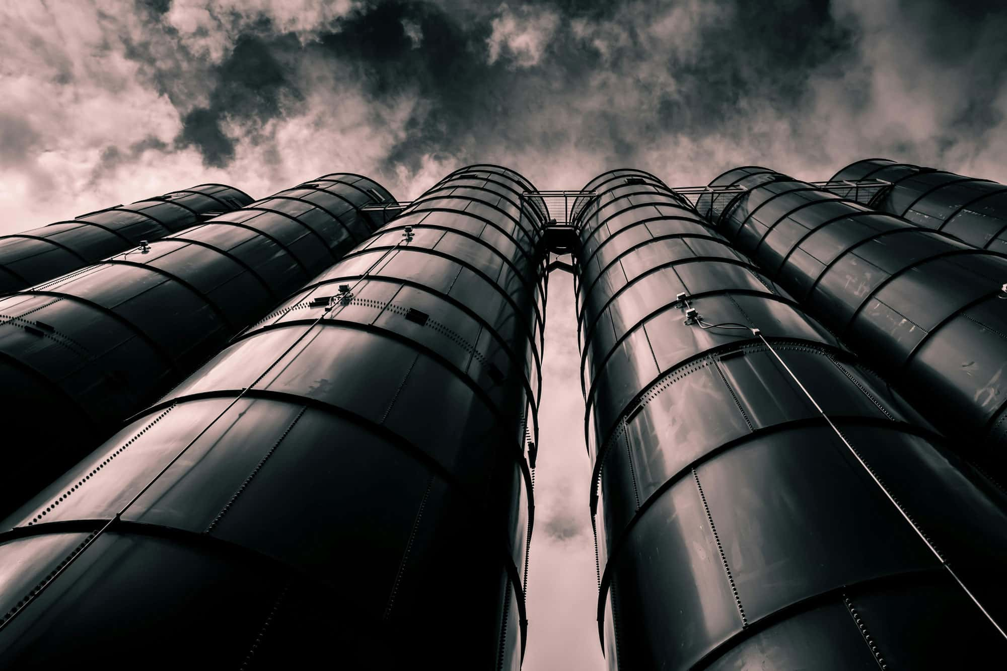 Mehrere hohe, schwarze Silos ragen in den Himmel. Die Perspektive von unten betont die Höhe der Strukturen. Der Himmel ist wolkig und dramatisch.