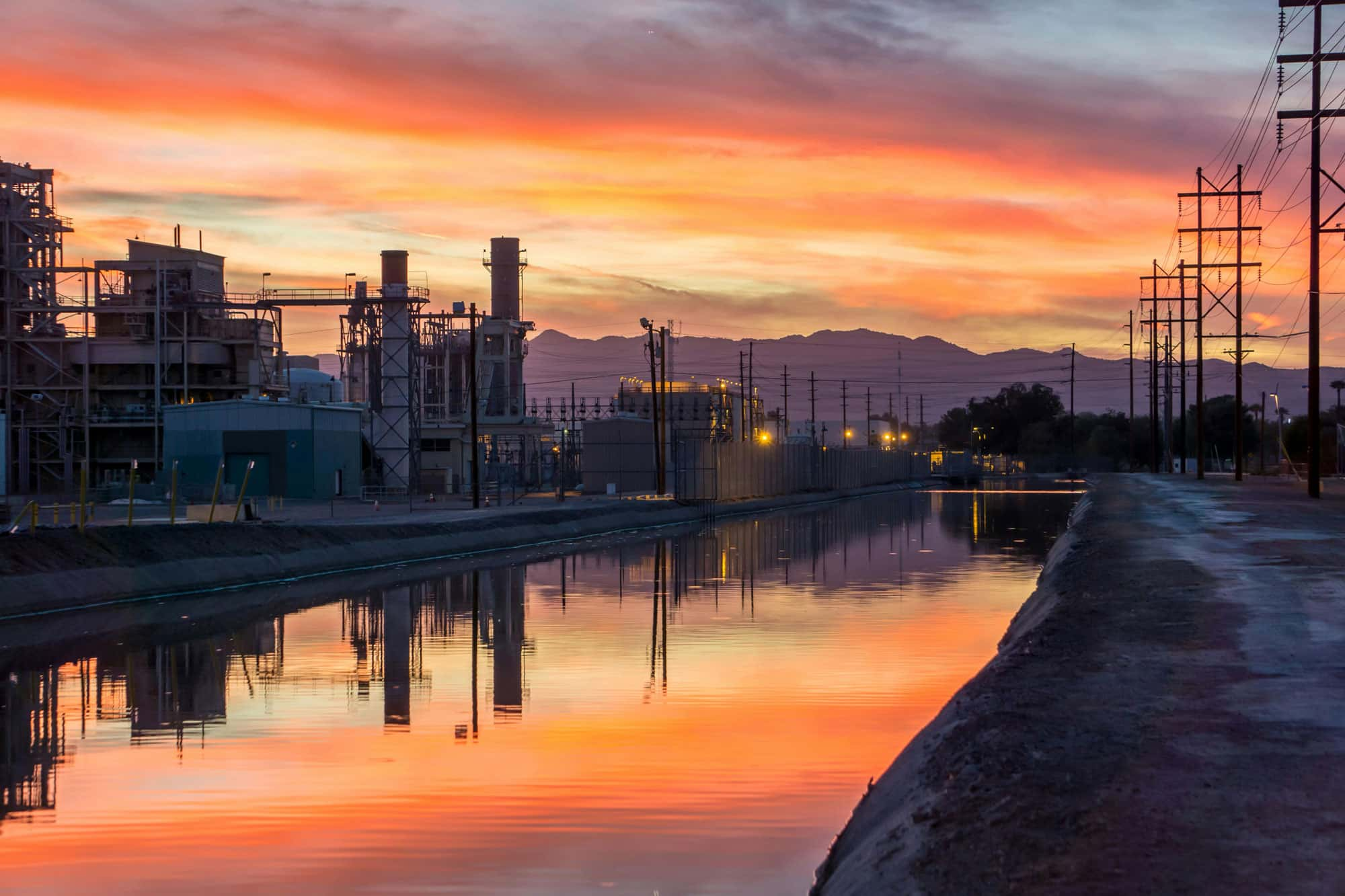 Eine Industrieanlage bei Sonnenuntergang, mit Gebäuden und Schornsteinen, die sich im ruhigen Wasser eines Kanals spiegeln. Der Himmel ist in warmen Farbtönen gefärbt, und im Hintergrund sind Berge sichtbar.