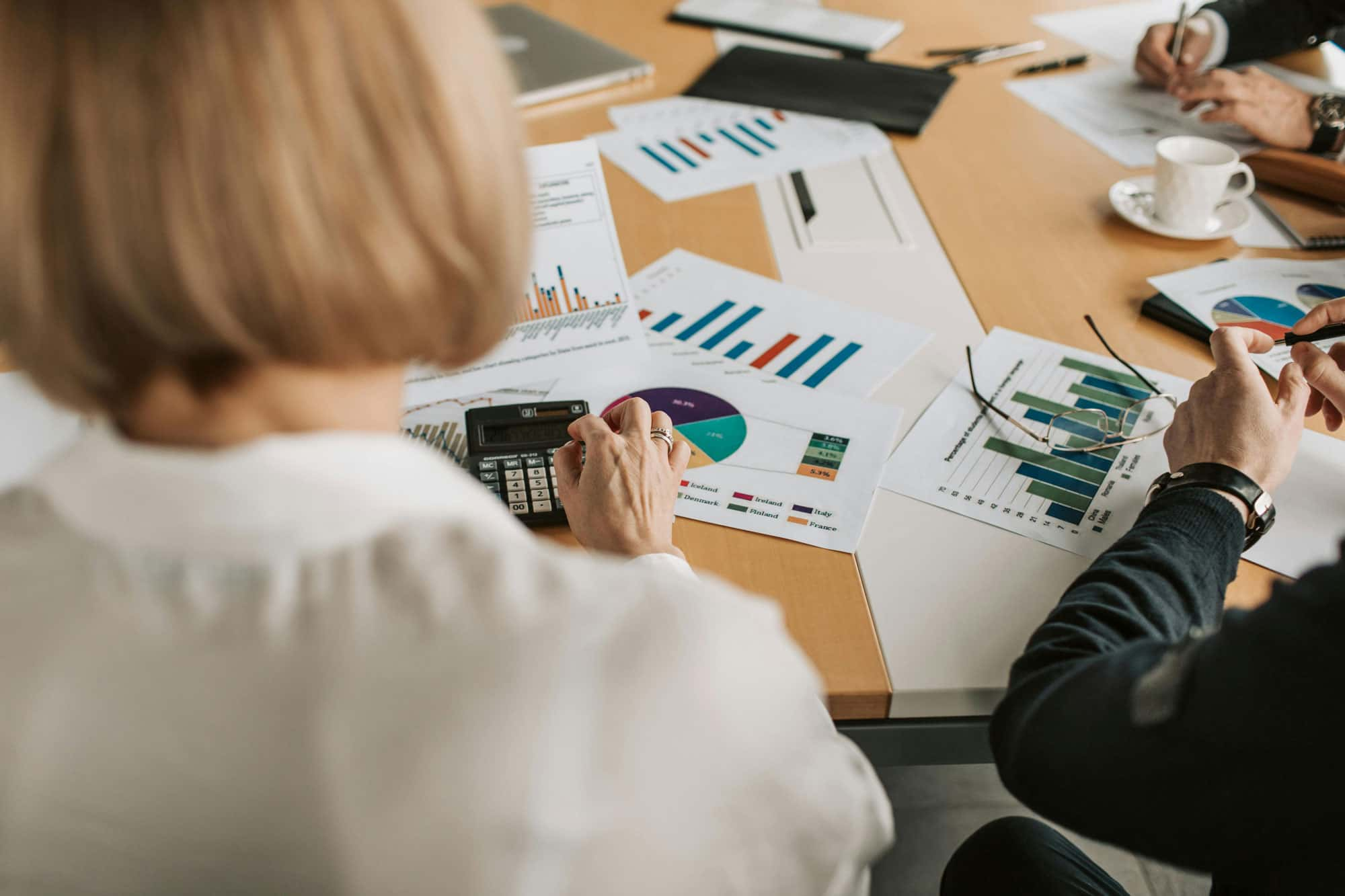 Personen sitzen an einem Tisch und analysieren verschiedene Diagramme und Berichte. Eine Person verwendet einen Taschenrechner, während andere Notizen machen und die Dokumente besprechen.