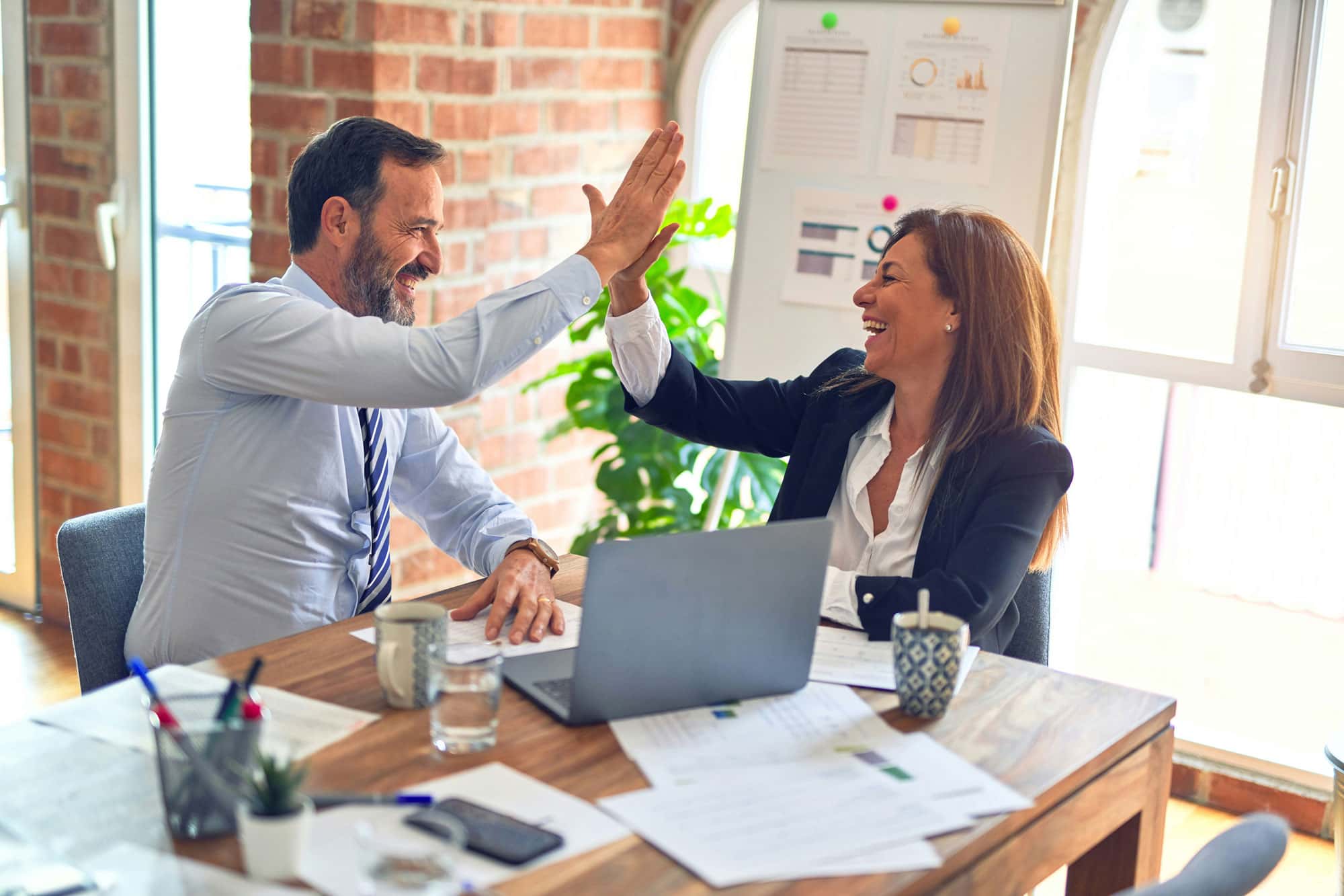 Ein Mann und eine Frau im Büro geben sich ein High-Five, während sie an einem Tisch mit einem Laptop, Dokumenten und Kaffeetassen sitzen. Sie lachen und feiern offenbar einen gemeinsamen Erfolg.