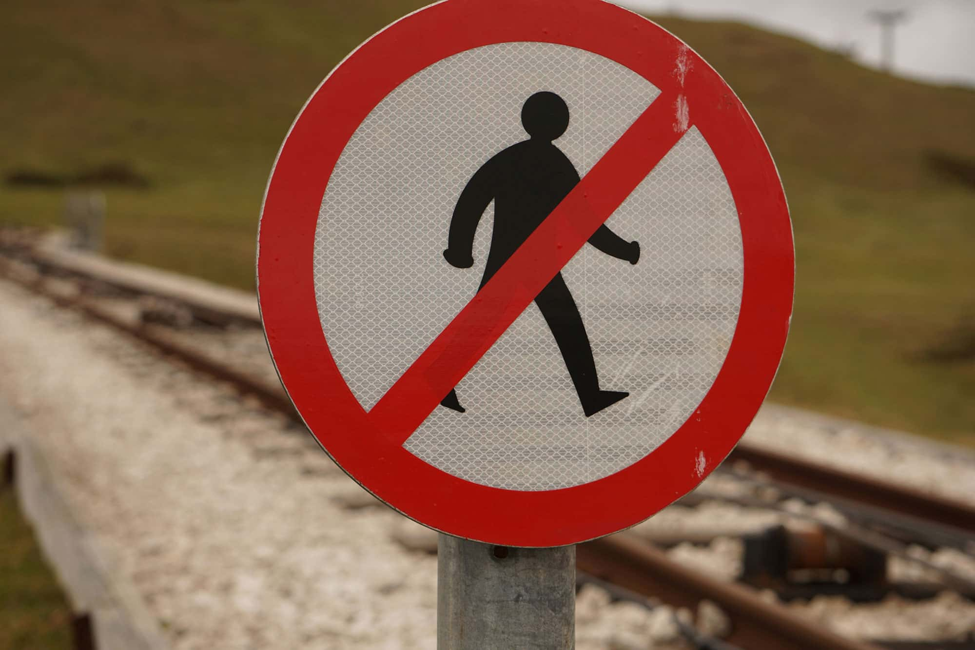 Ein rundes Verbotsschild mit einem durchgestrichenen Fußgängersymbol steht neben einem Bahngleis und warnt davor, die Gleise zu betreten. Im Hintergrund sind Schienen und ein grasbewachsener Hügel zu sehen.