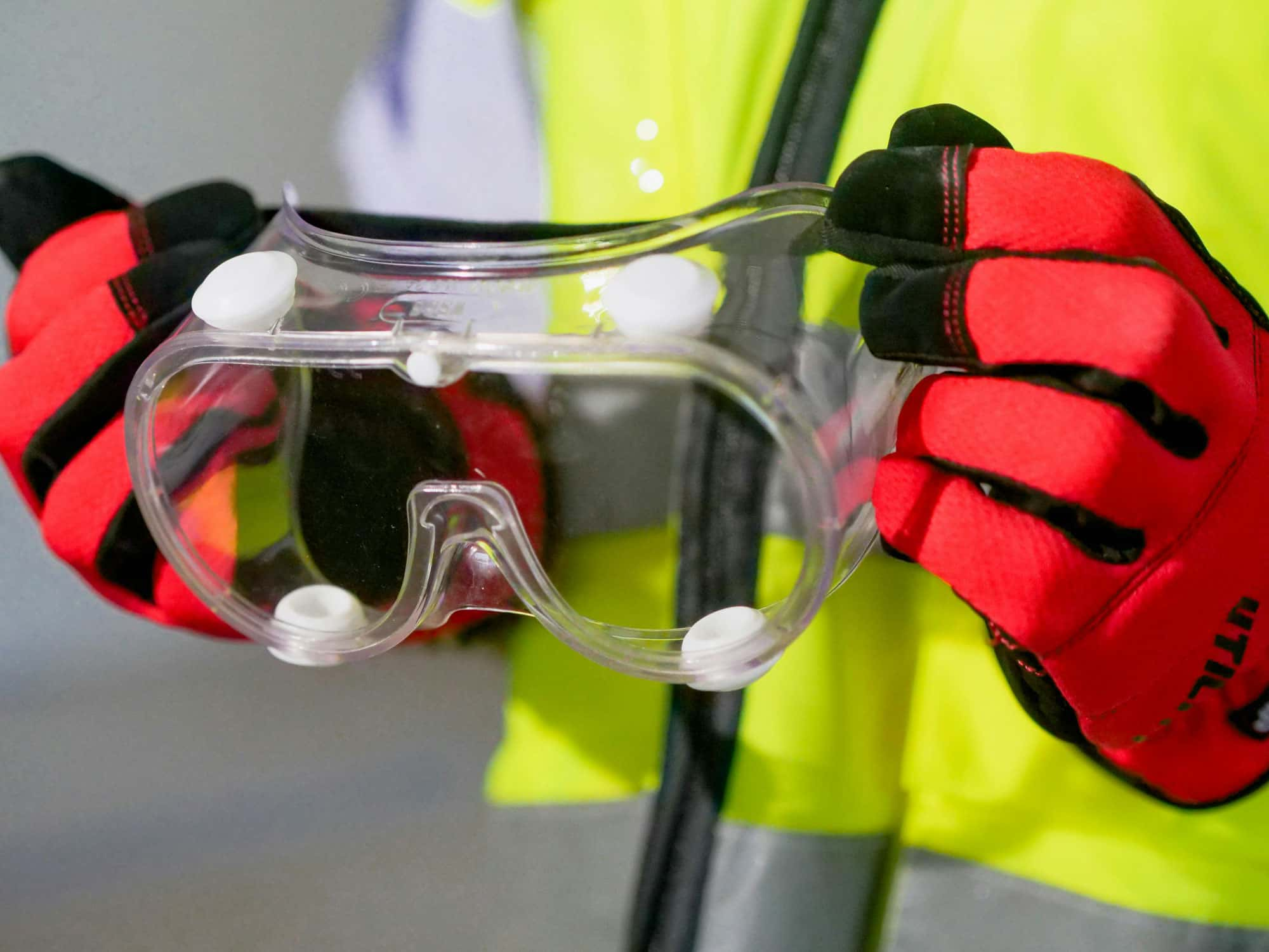 Person in Schutzkleidung hält eine Schutzbrille in rot-schwarzen Handschuhen.