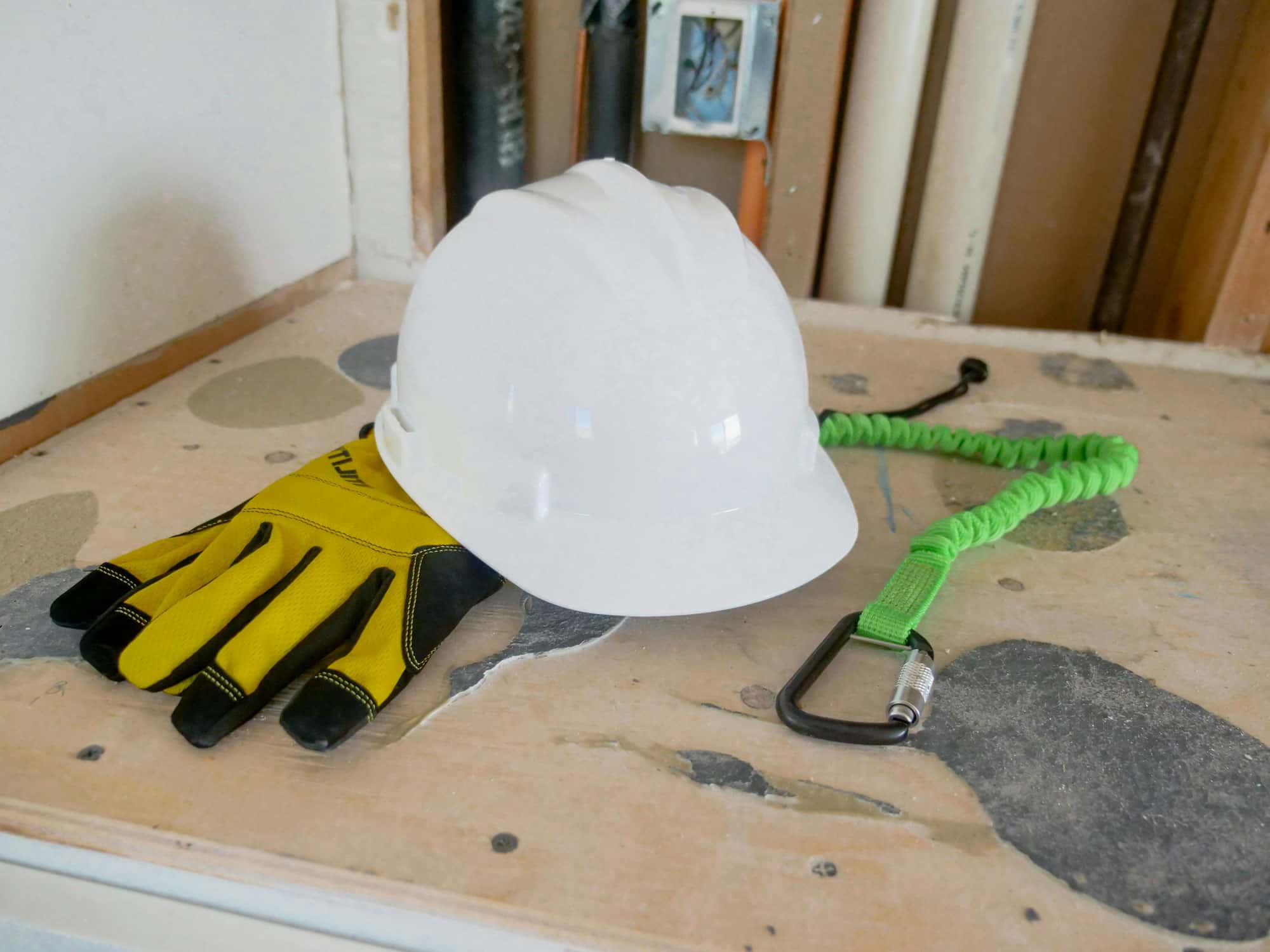 Schutzausrüstung auf einem Tisch: weißer Helm, gelbe Handschuhe und grüne Sicherungsleine mit Karabiner.