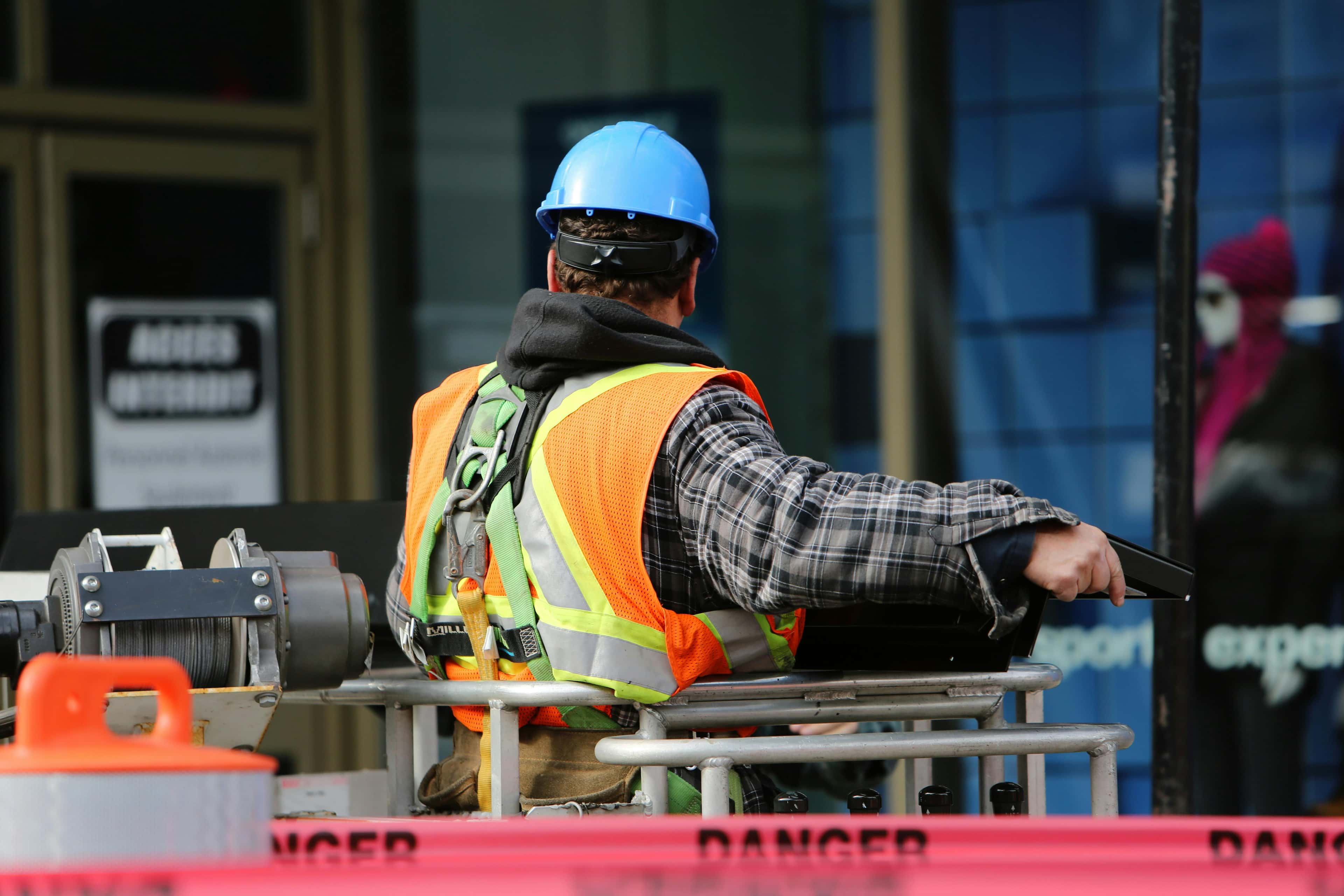 Ein Bauarbeiter trägt einen blauen Schutzhelm und eine orangefarbene Warnweste. Er sitzt auf einer Hebebühne und arbeitet an einer Baustelle.