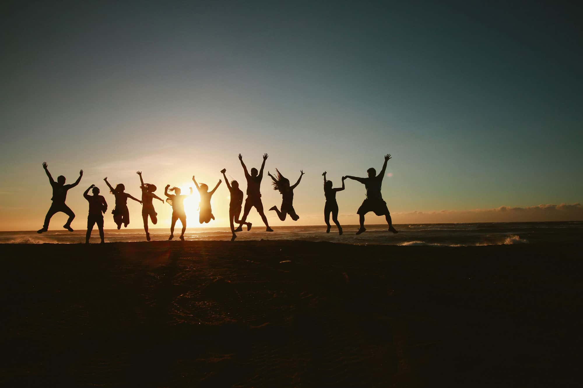 Gruppe von Menschen springt vor Freude am Strand bei Sonnenuntergang, Silhouetten vor einem farbenfrohen Himmel. 