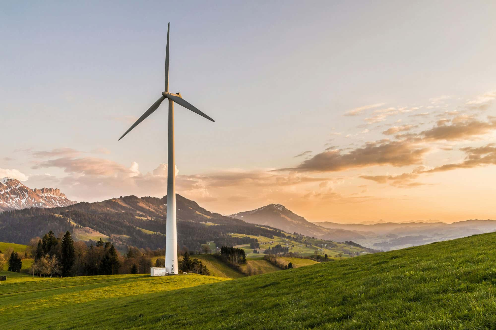 Windkraftanlage auf einem grünen Hügel bei Sonnenuntergang, mit malerischen Bergen im Hintergrund.