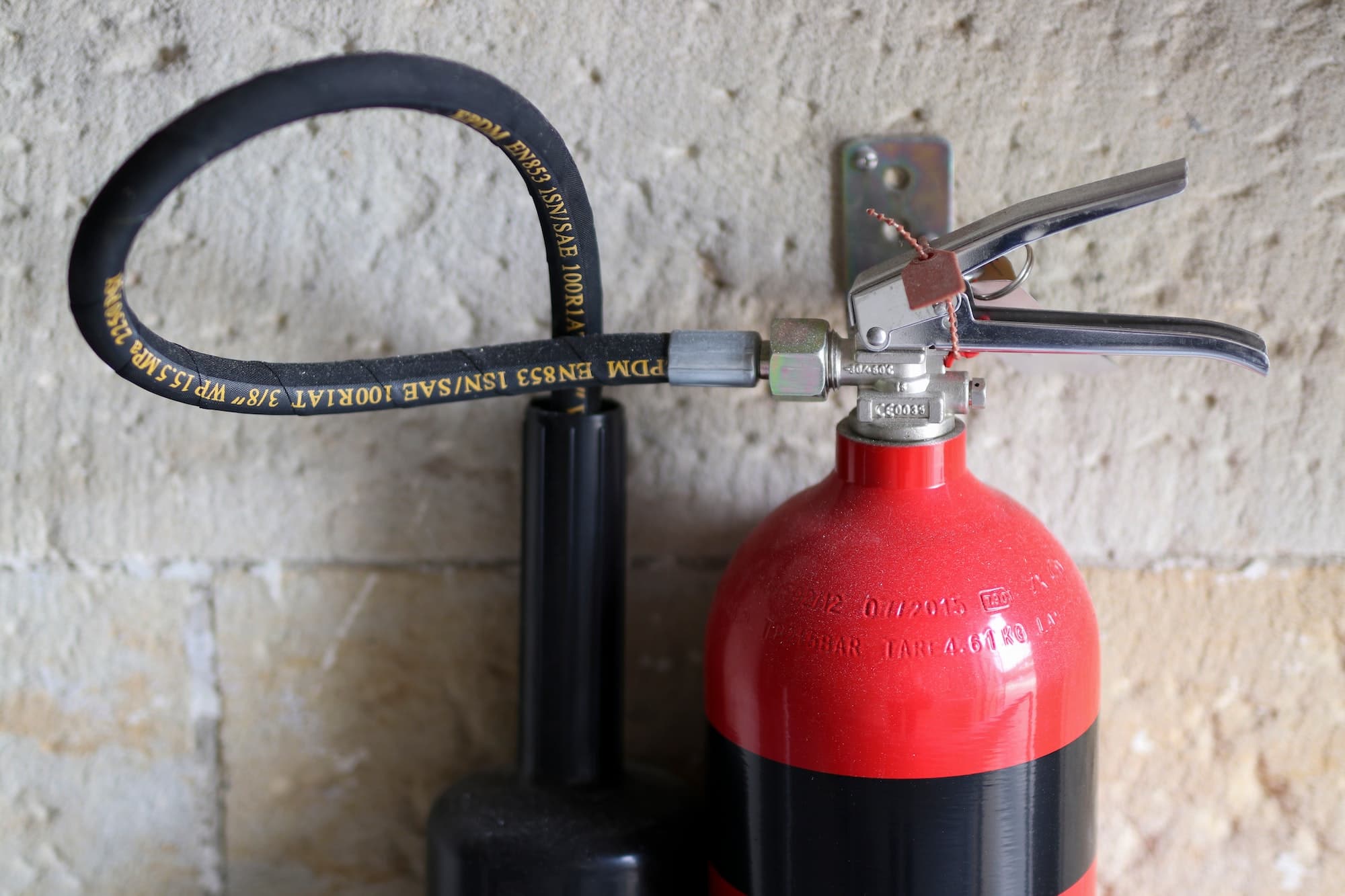 Ein roter Feuerlöscher ist an einer Wand befestigt. Der schwarze Schlauch und der silberne Griff sind deutlich sichtbar. Der Hintergrund besteht aus einer hellen, strukturierten Wand.