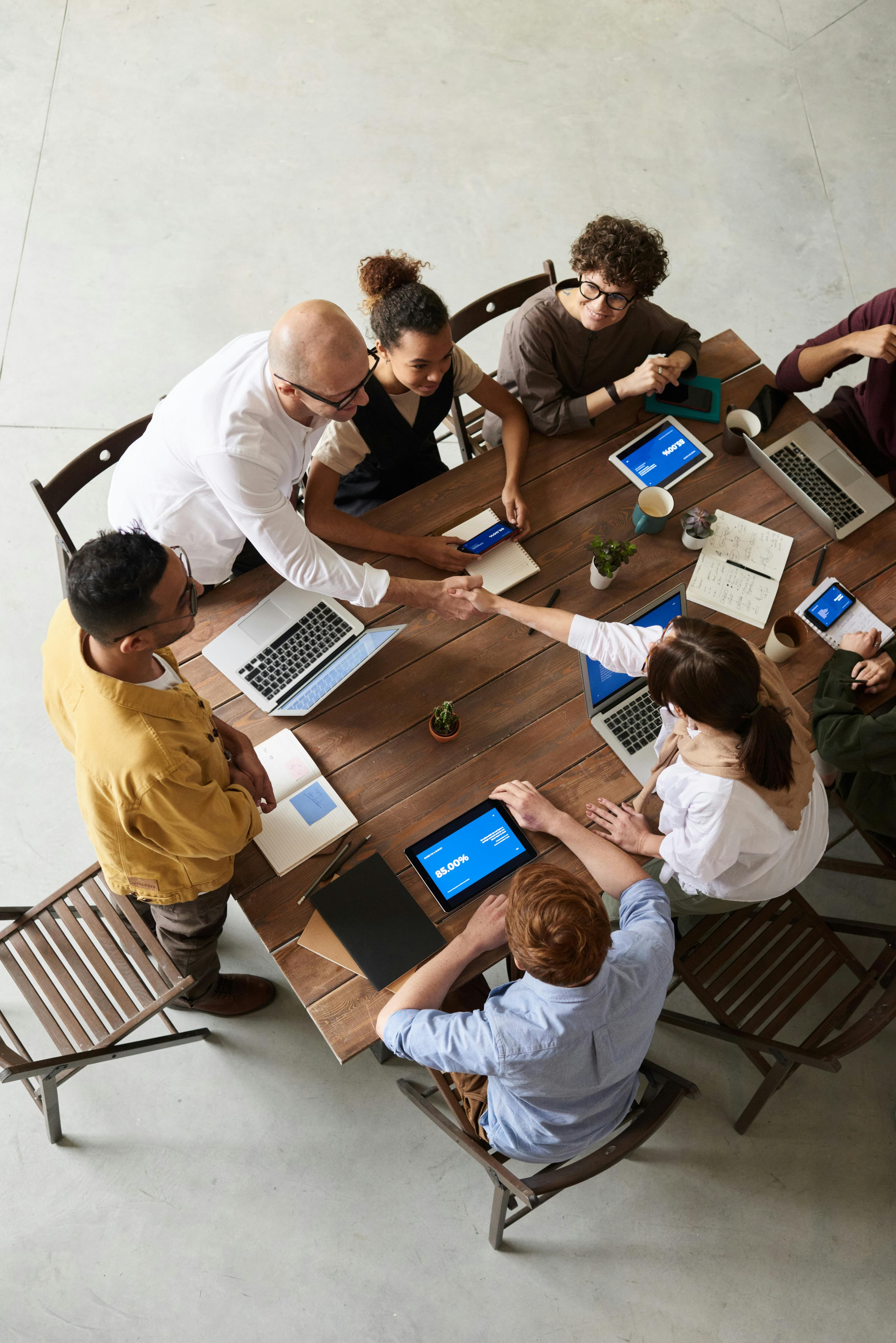 Eine Gruppe von Menschen sitzt um einen Tisch, diskutiert und arbeitet gemeinsam an Laptops und Tablets. Zwei Personen schütteln sich zur Begrüßung die Hände.