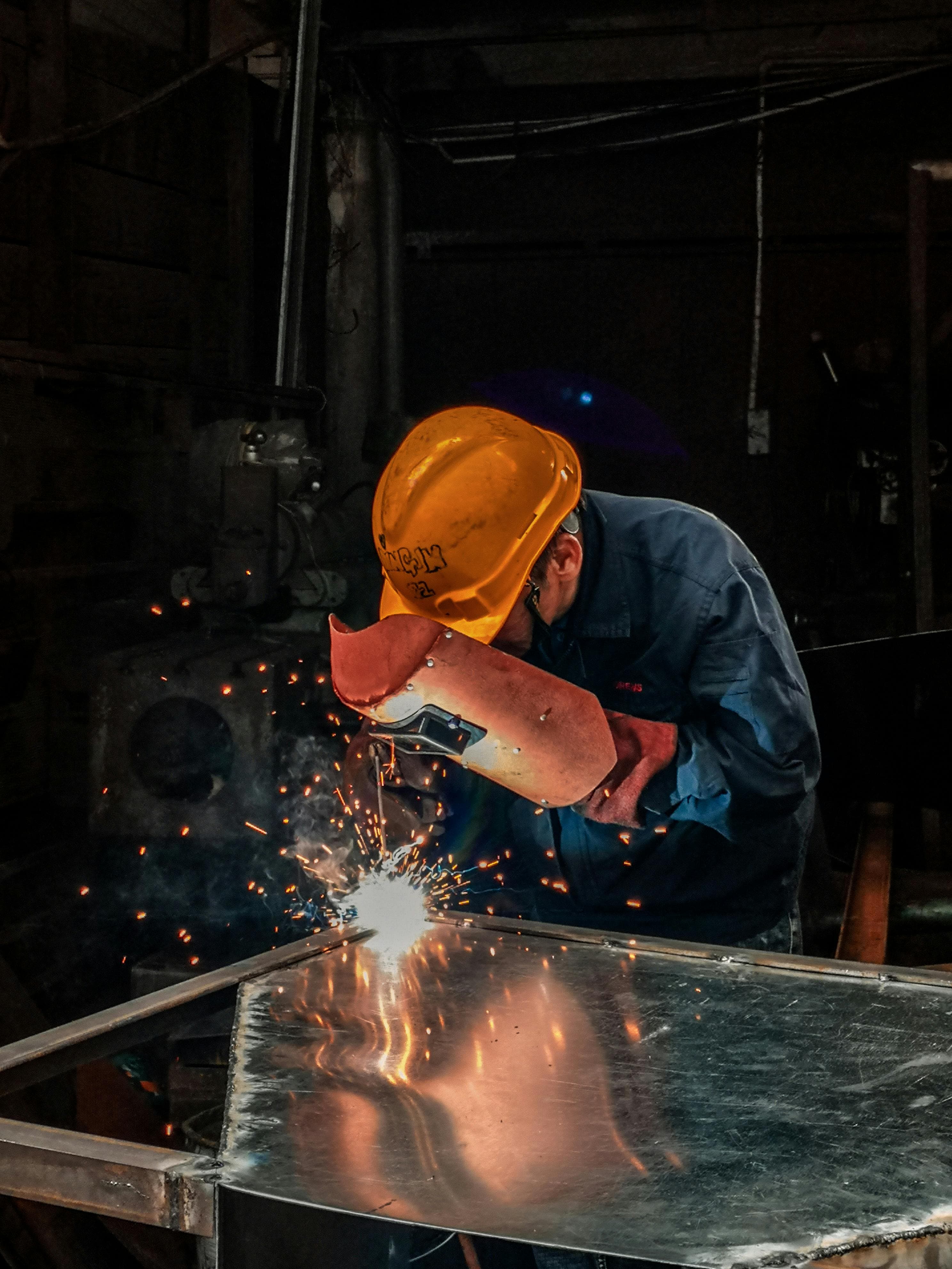 Ein Arbeiter trägt einen gelben Schutzhelm und eine Schweißmaske und schweißt an einem Metallrahmen in einer Werkstatt, wobei Funken sprühen.