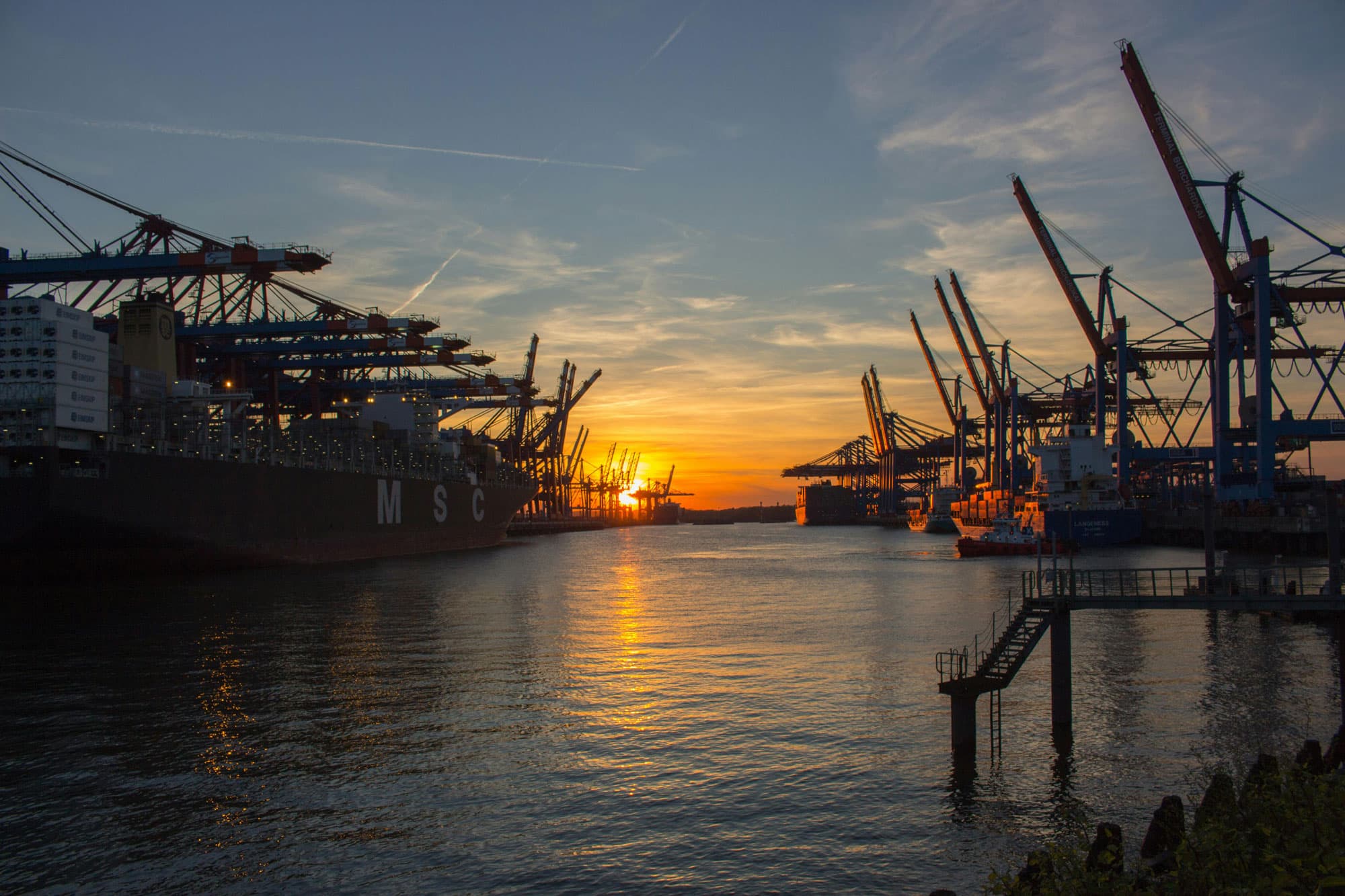 Containerhafen bei Sonnenuntergang, große Frachtschiffe und Kräne am Wasser, spiegelnde Lichtreflexionen auf der Wasseroberfläche.