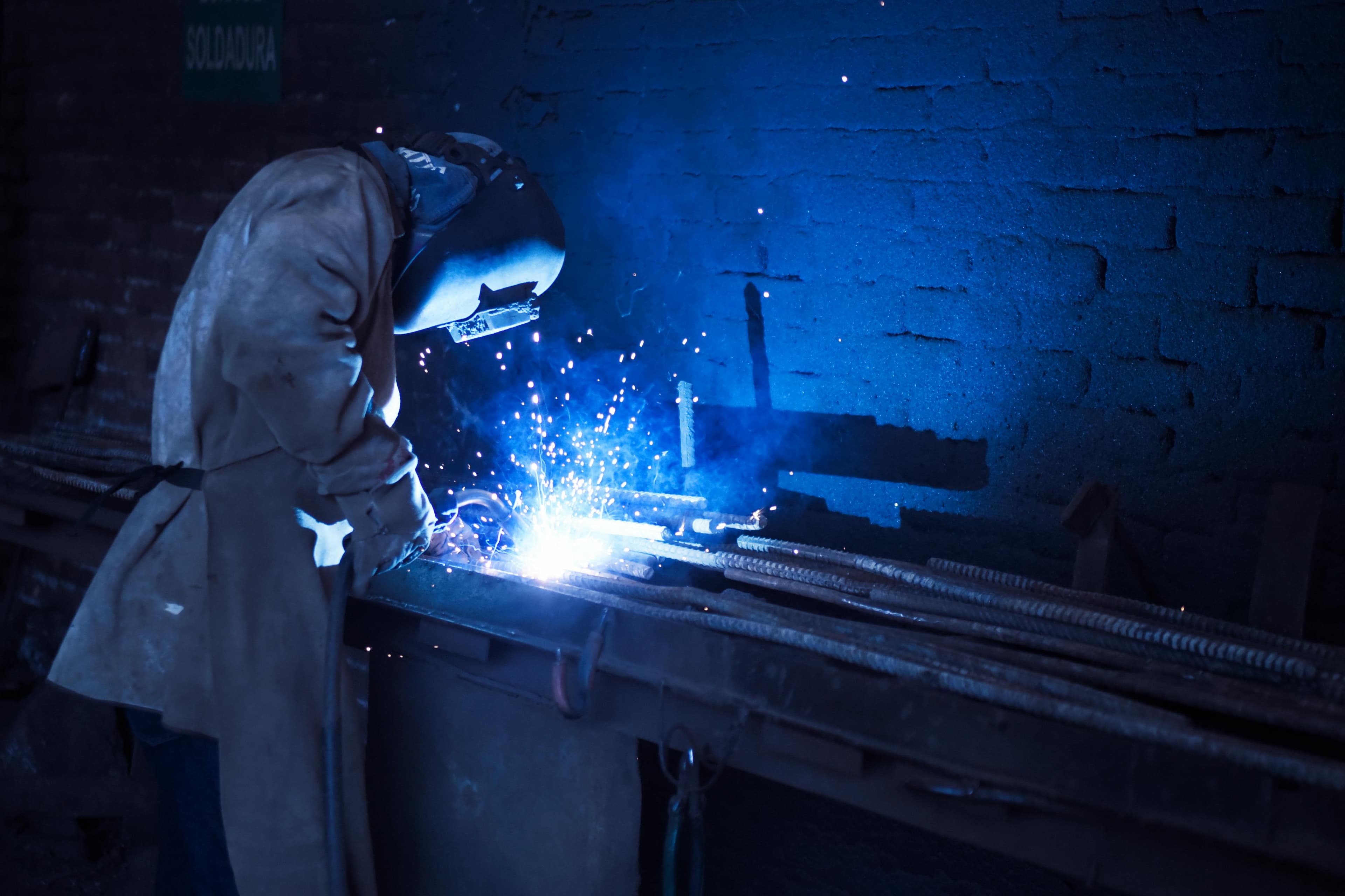 Ein Schweißer in Schutzkleidung und Helm, der in einer dunklen Werkstatt arbeitet. Blaue Funken und Lichtblitze erhellen den Bereich um den Schweißer.