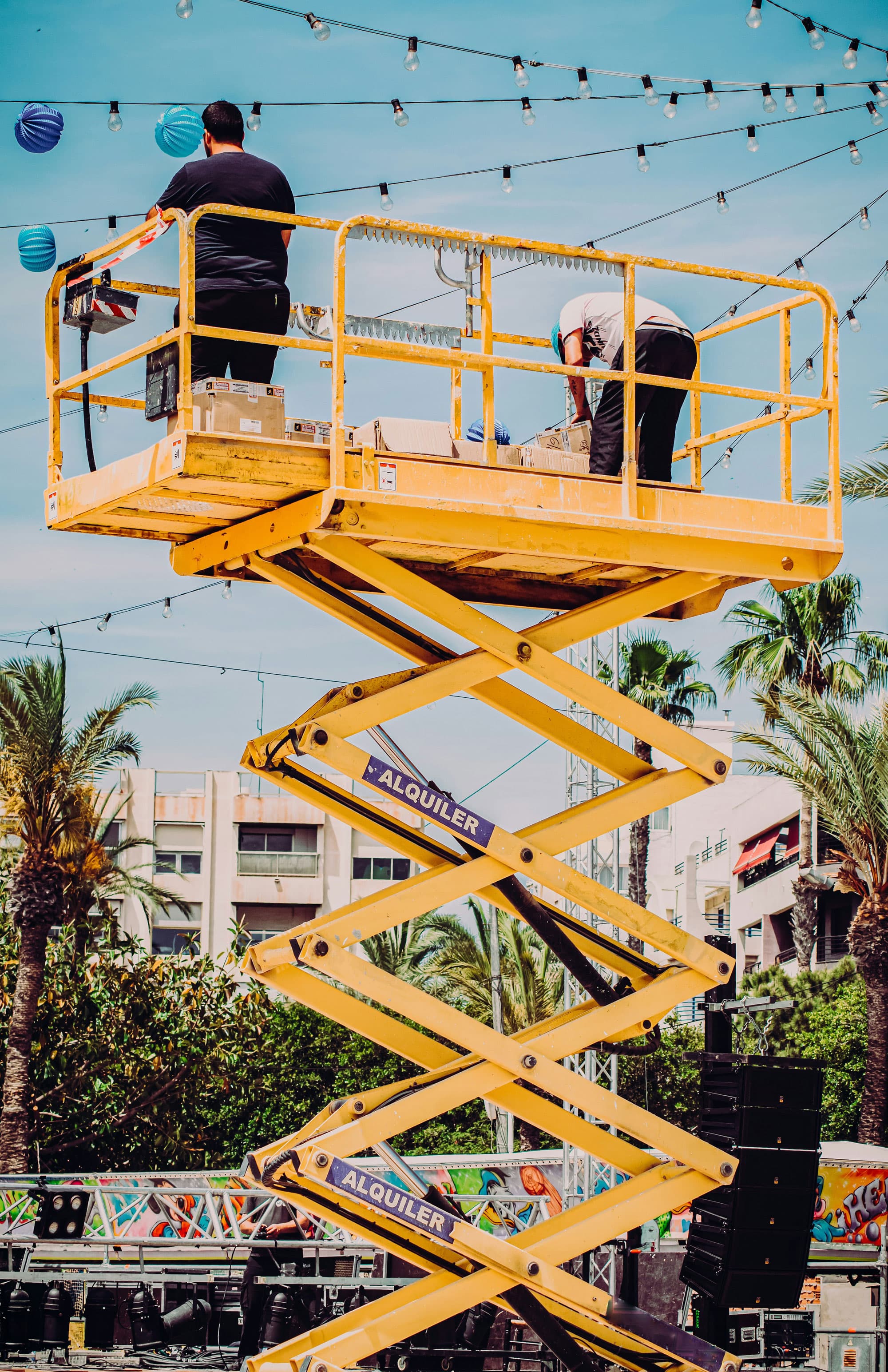 Zwei Arbeiter auf einer gelben Hebebühne im Freien, umgeben von Palmen und hängenden Lichtern, beim Aufbau oder der Inspektion von Equipment.