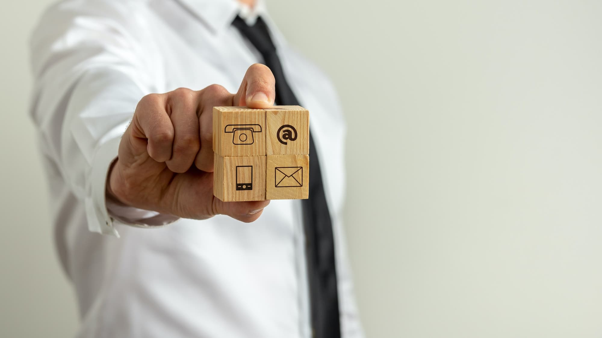 Eine Person in einem weißen Hemd und schwarzer Krawatte hält Holzklötze mit Symbolen für Telefon, E-Mail, Handy und Post in der Hand. Die Symbole stehen für Kontaktmöglichkeiten.