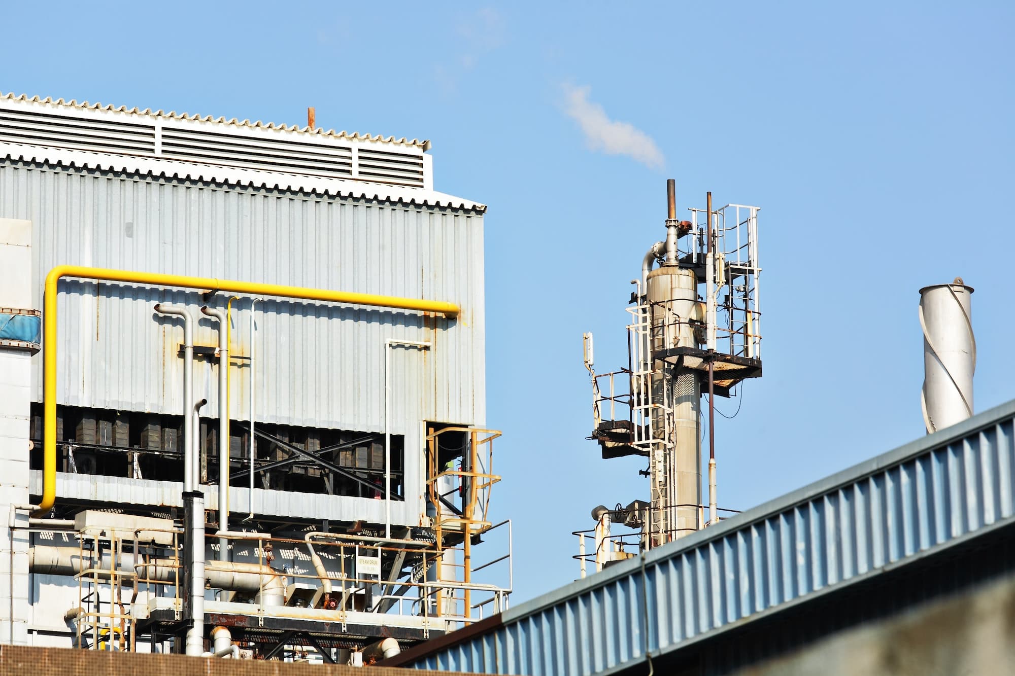 Ein Industriegebäude mit gelben Rohren und einem Schornstein, der Dampf abgibt. Im Hintergrund ist ein klarer blauer Himmel zu sehen. Das Bild zeigt eine industrielle Anlage.