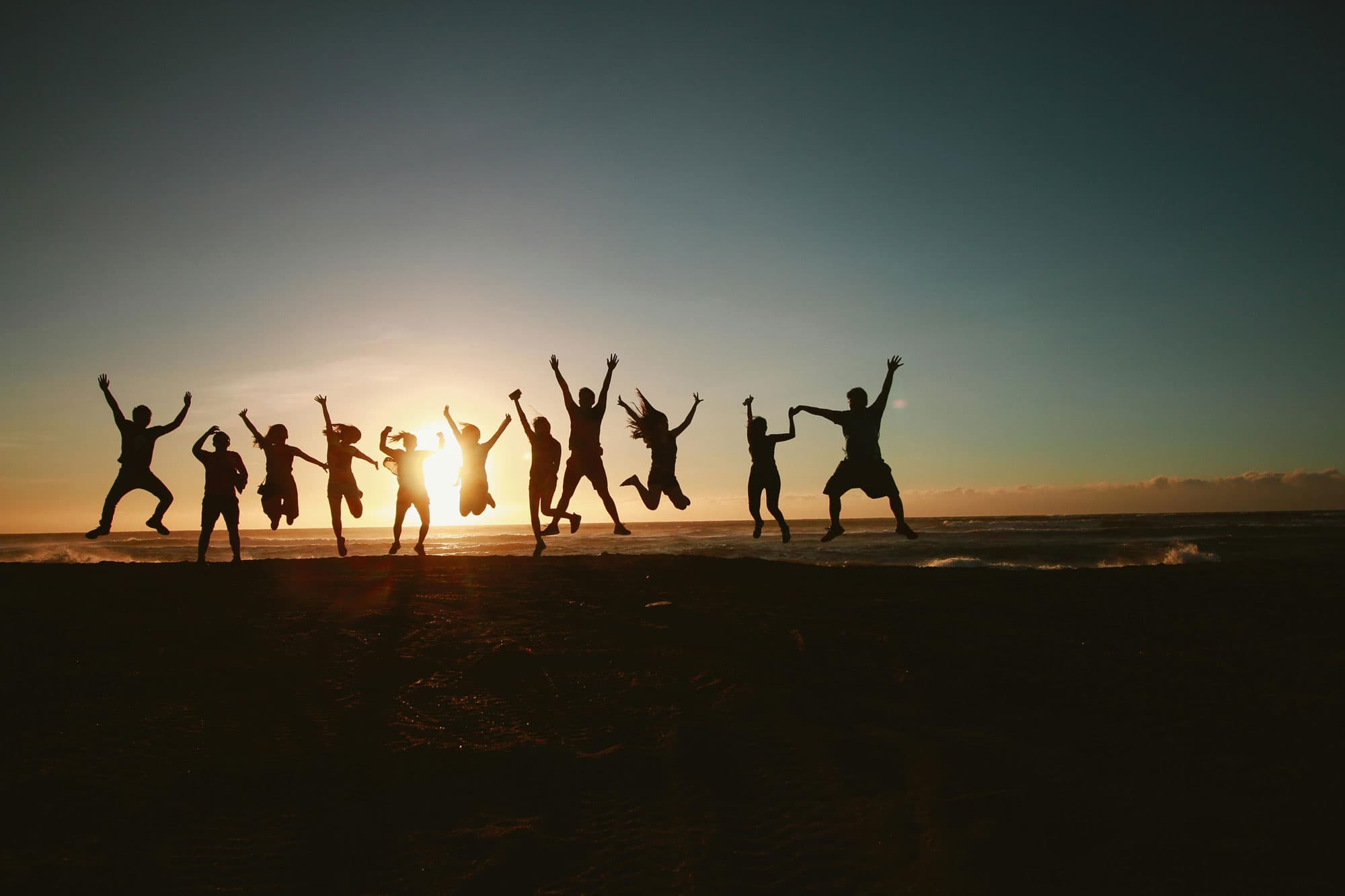 Gruppe von Menschen springt vor Freude am Strand bei Sonnenuntergang, Silhouetten vor einem farbenfrohen Himmel.