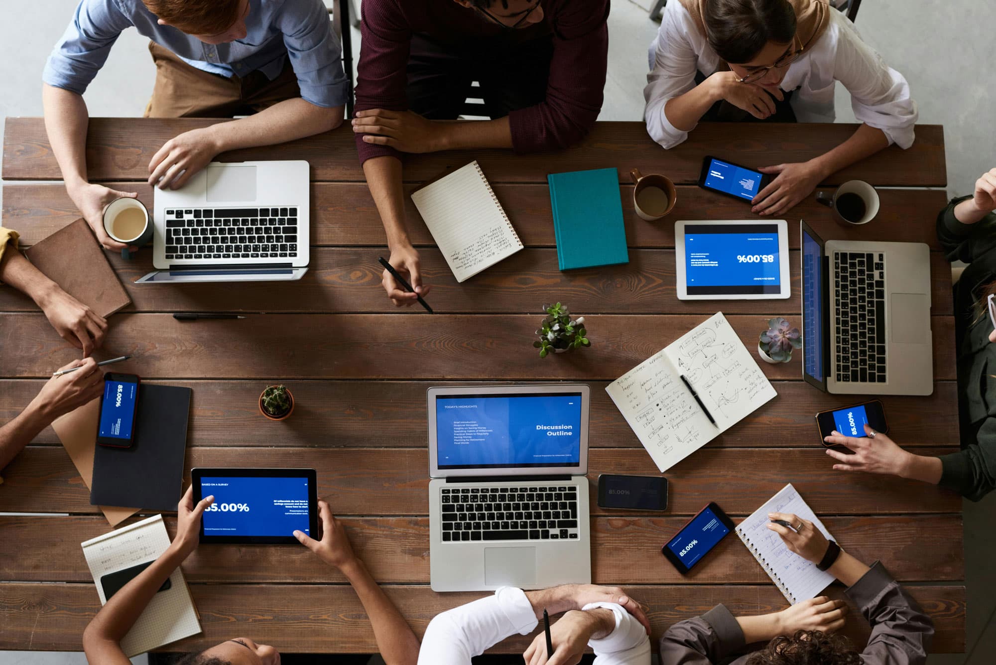 Team von Fachleuten arbeitet zusammen an einem Holztisch, Laptops, Tablets und Notizbücher vor sich, während sie an einem gemeinsamen Projekt arbeiten.